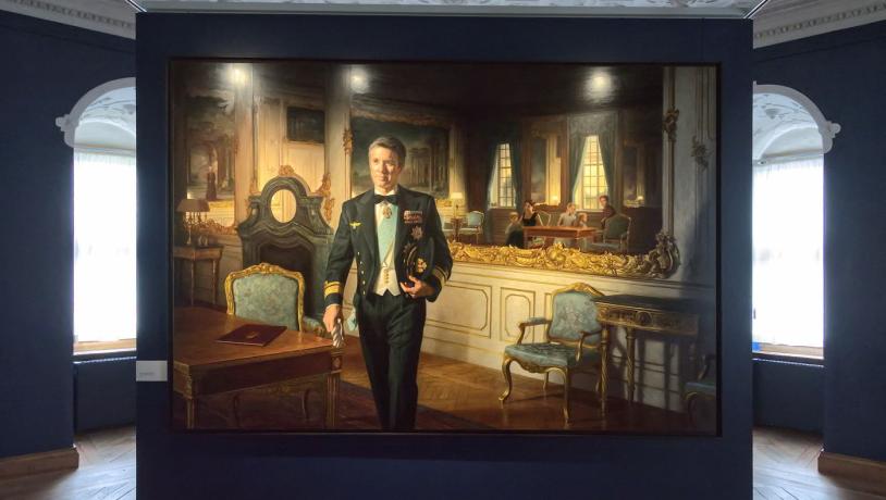 Painting of Crown Prince Frederik in a suit and decorations, hanging on a dark blue wall with spotlight.